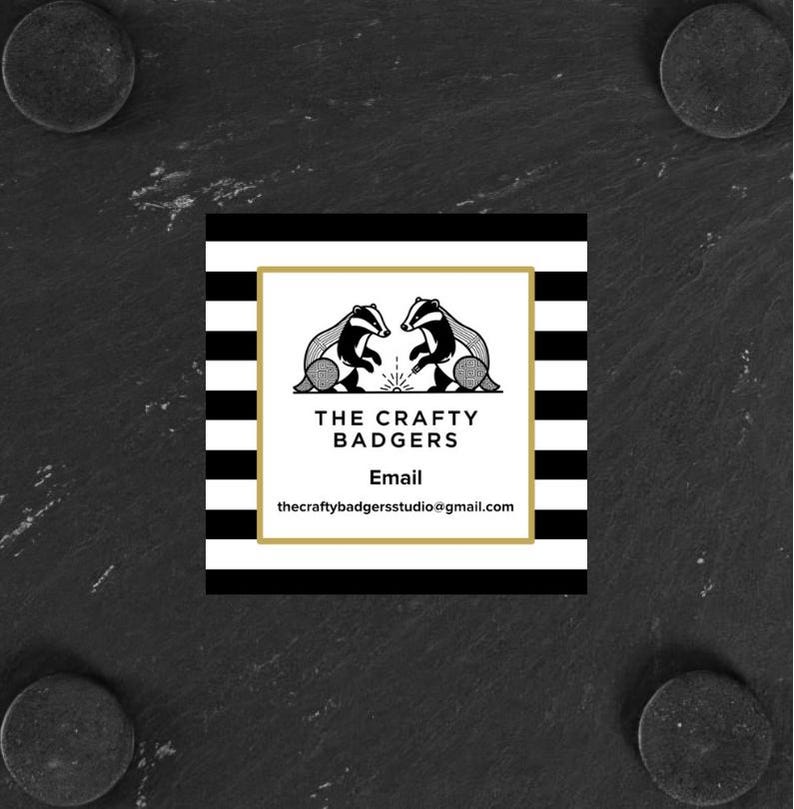 May include: A square business card featuring a black and white horizontal striped border. The centre white panel displays a gold frame, two badger illustrations, and the text: THE CRAFTY BADGERS, Email, thecraftybadgersstudio@gmail.com.