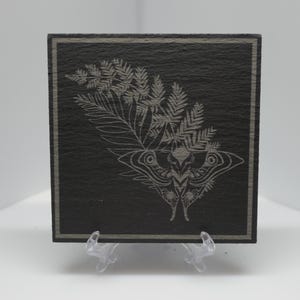 May include: Square black slate coaster featuring an etched design of a moth and fern leaves. The coaster has a thin, lighter border and is displayed on a clear plastic stand. The coaster measures approximately 10 cm x 10 cm.