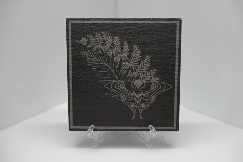 May include: A square black slate coaster featuring a laser-engraved design of a moth positioned beneath a fern frond. The coaster has a thin grey border etched around the perimeter and is displayed on a clear plastic stand against a white background.