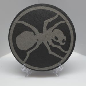 May include: A circular black slate coaster featuring a grey engraved graphic of an ant centred within a circular border. The coaster is supported by a small, clear plastic display stand, showcasing the textured surface and the insect design.
