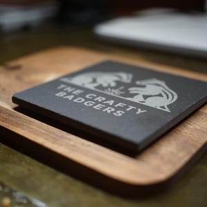 May include: A square black slate coaster featuring a white engraved graphic of two badgers facing each other and the text "THE CRAFTY BADGERS" positioned on a rectangular wooden board with rounded corners and a visible wood grain texture.