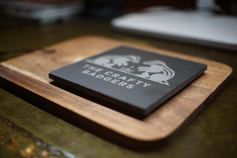 May include: A square black slate coaster featuring a white engraved graphic of two badgers facing each other and the text "THE CRAFTY BADGERS" positioned on a rectangular wooden board with rounded corners and a visible wood grain texture.