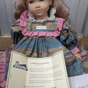 May include: A Jesmar doll named Veronique, wearing a plaid dress with pink accents and a white lace collar. The doll has auburn hair and is holding a certificate of authenticity. The doll is 20 inches tall.