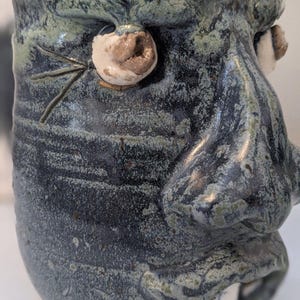 Könnte beinhalten: Eine handgefertigte Keramik-Tasse mit Gesichtsdesign. Die Tasse ist überwiegend dunkelblau mit grünen und braunen Akzenten. Sie hat hervorstehende Augen, eine Nase und einen Mund mit einem einzelnen Zahn. Die Tasse hat eine strukturierte Oberfläche und ein einzigartiges, künstlerisches Aussehen.