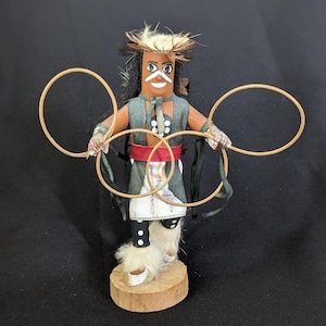 Può includere: Una bambola Kachina nativa americana con corpo in legno, indossa un gilet verde, una gonna bianca e una cintura rossa. La bambola ha un viso dipinto, capelli di paglia e tiene tre cerchi di legno. Si trova su una base di legno.