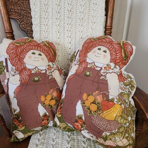 May include: Two decorative pillows featuring a vintage doll design. The dolls have red hair, brown overalls, and are holding baskets of vegetables. The pillows are on a wooden rocking chair with a cream-colored knitted throw.