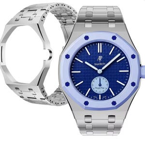 Könnte beinhalten: Eine silberfarbene Armbanduhr mit blauem, strukturiertem Zifferblatt, das den Schriftzug AP x Swatch und Royal Pop trägt. Die Uhr hat eine hellblaue achteckige Lünette mit dunkelblauen Schrauben, silberne Zeiger und ein silbernes Gliederarmband.