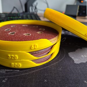 May include: A set of yellow sanding discs with grit levels 60, 320, and 800, stacked in a yellow holder. A separate yellow sanding block with "40" printed on it is nearby. The discs are reddish-brown. Measurements are in metric.