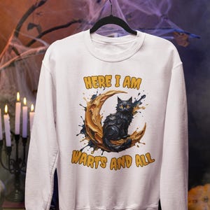 May include: A white crewneck sweatshirt featuring a graphic print of a black cat sitting on a crescent moon. The text "HERE I AM" is printed above the moon and "WARTS AND ALL" is printed below the moon in a bold, orange, rounded font.