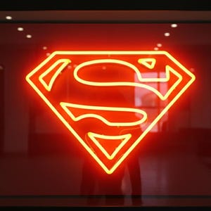 Può includere: Un'insegna al neon con il logo di Superman, un diamante giallo con una "S" gialla all'interno. L'insegna si illumina brillantemente su uno sfondo rosso, racchiusa in una cornice nera. L'immagine è un primo piano dell'iconico simbolo.