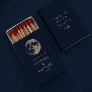May include: Two dark navy blue matchboxes with red-tipped matches. One box is open, displaying a moon graphic, the text "TO THE MOON AND BACK", "KENDALL & MICHAEL", and "08.16.2027". The closed box features "I LOVE YOU BEYOND THE STARS" and an infinity symbol.