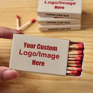 May include: A rectangular white matchbox with red text that reads Your Custom Logo/Image Here. The box is partially open, revealing wooden matches with red tips. Additional stacked matchboxes are visible, each printed with custom your text.