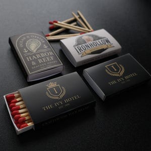 May include: Four rectangular matchboxes with red-tipped wooden matches. Two black boxes feature gold text for The Ivy Hotel, one black box displays Harbor & Reef Seafood House branding, and one white box shows the IronHollow Brewing Co logo.