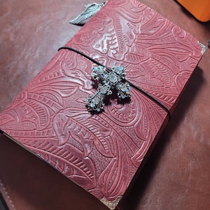 May include: A burgundy leather journal with an embossed floral design. The journal is secured with a black elastic band and features a decorative jeweled cross and silver wing charm. The corners are protected with gold-colored metal.