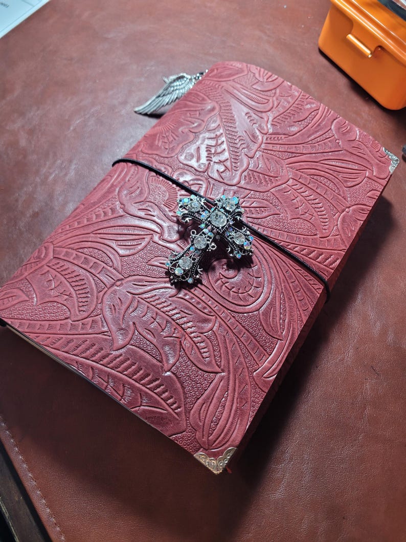May include: A burgundy leather journal with an embossed floral design. The journal is secured with a black elastic band and features a decorative jeweled cross and silver wing charm. The corners are protected with gold-colored metal.