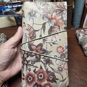 May include: A rectangular leather journal cover featuring a beige background with a printed floral pattern in shades of brown, rust, and cream. The cover is secured by a thin, sage green elastic cord wrapped diagonally around the exterior.