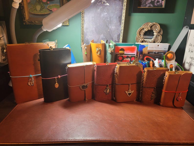 May include: A collection of leather journals in various colors, including brown, black, and red. Each journal features a clasp closure and decorative charms. Some have elastic closures. The journals are arranged on a brown surface.