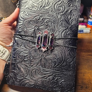 May include: Black leather journal with embossed floral design. A decorative clasp with three purple gems and a red border secures the journal. The journal is held together with a black elastic band.