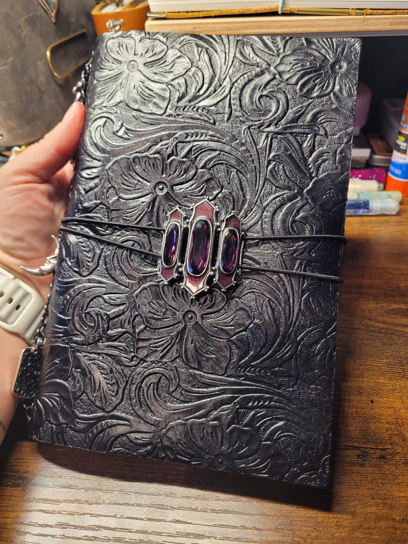 May include: Black leather journal with embossed floral design. A decorative clasp with three purple gems and a red border secures the journal. The journal is held together with a black elastic band.