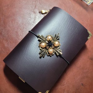 May include: A dark burgundy leather journal with a decorative gold and jewel embellishment. The journal features a black elastic closure and gold corner protectors. The embellishment includes pearls and a central dark blue stone.