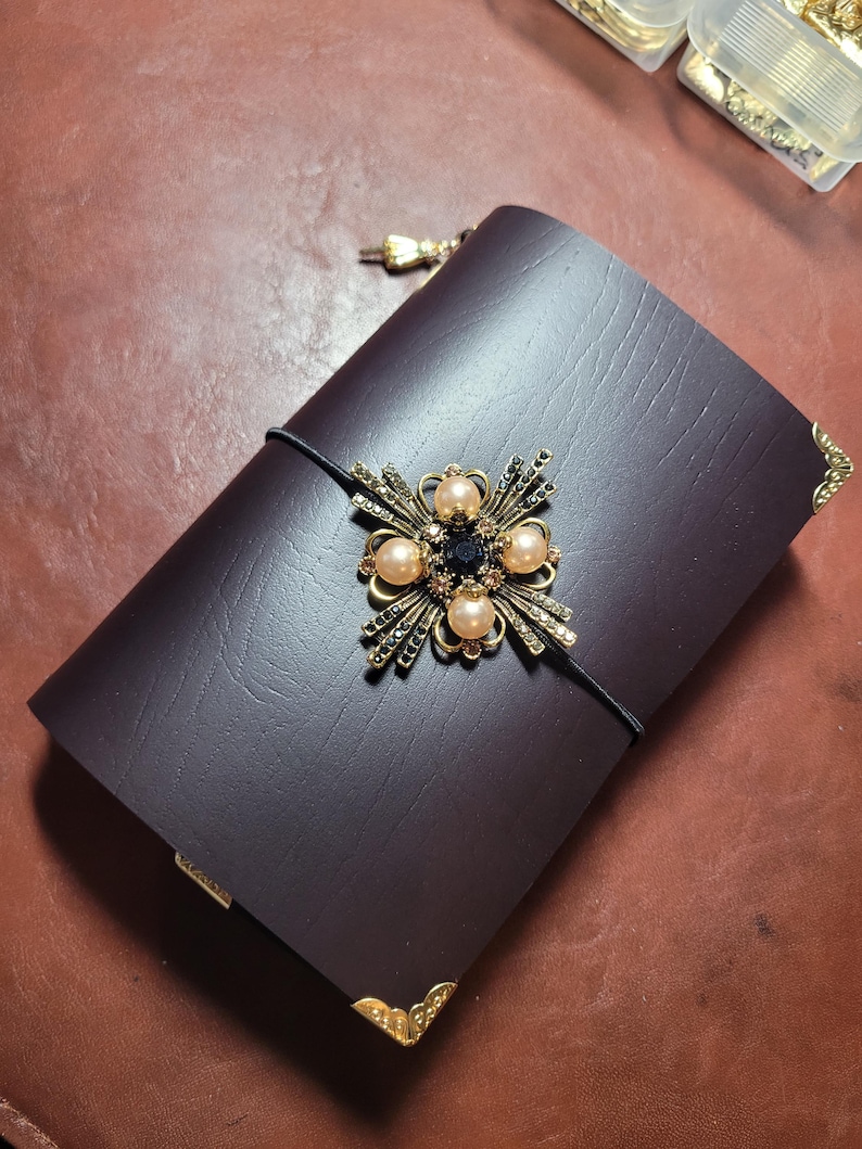May include: A dark burgundy leather journal with a decorative gold and jewel embellishment. The journal features a black elastic closure and gold corner protectors. The embellishment includes pearls and a central dark blue stone.