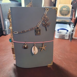 May include: A gray leather journal with gold corner protectors and a pink elastic closure. The journal is adorned with gold charms, including a seashell, starfish, and a rose. A gold chain with a small locket and a dragonfly charm is also attached.