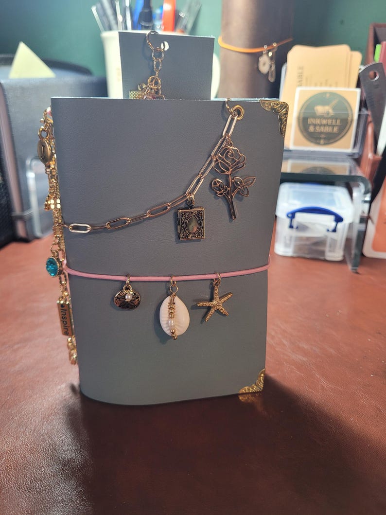 May include: A gray leather journal with gold corner protectors and a pink elastic closure. The journal is adorned with gold charms, including a seashell, starfish, and a rose. A gold chain with a small locket and a dragonfly charm is also attached.
