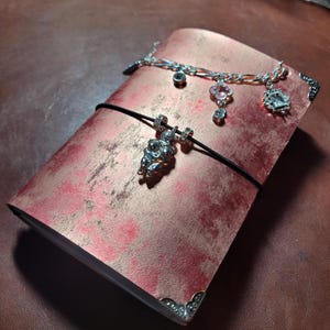 May include: A red and gold journal with a black elastic closure and silver corner protectors. A silver charm bracelet with various charms is draped across the journal. A silver charm hangs from the elastic.