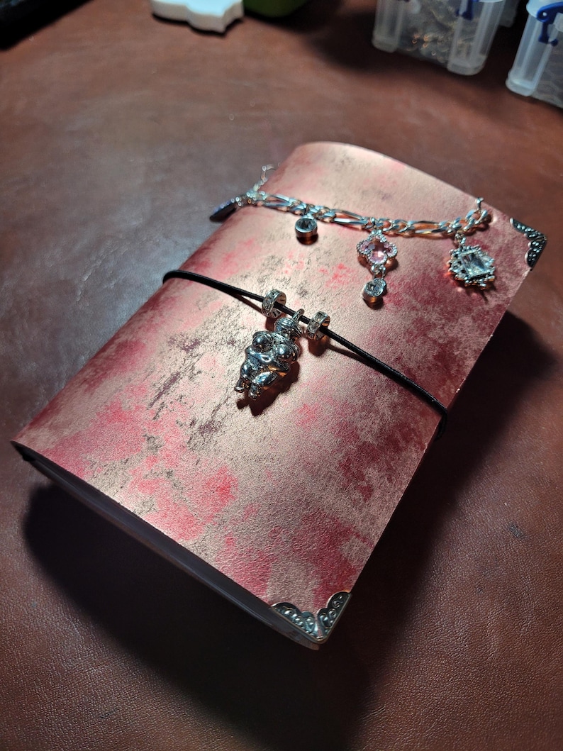 May include: A red and gold journal with a black elastic closure and silver corner protectors. A silver charm bracelet with various charms is draped across the journal. A silver charm hangs from the elastic.