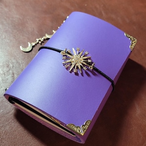 May include: A purple journal with gold corner protectors and a black elastic band. A gold starburst charm with clear crystals and a crescent moon charm dangle from the band. The journal is closed on a brown surface.