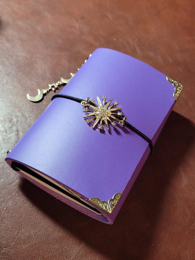 May include: A purple journal with gold corner protectors and a black elastic band. A gold starburst charm with clear crystals and a crescent moon charm dangle from the band. The journal is closed on a brown surface.