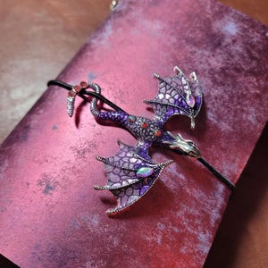 May include: A purple and silver dragon brooch with red and iridescent accents, resting on a textured, reddish-purple surface. The dragon has detailed wings and is attached to a black cord.