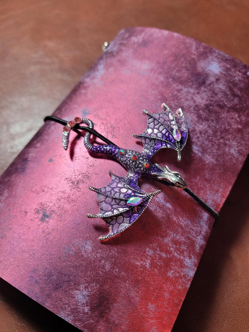May include: A purple and silver dragon brooch with red and iridescent accents, resting on a textured, reddish-purple surface. The dragon has detailed wings and is attached to a black cord.