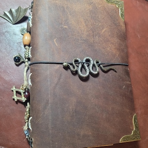 May include: A brown leather journal with decorative metal corner protectors and a snake-shaped clasp. A charm with a bat wing design and other decorative charms hang from the side. The journal is closed with a black elastic band.