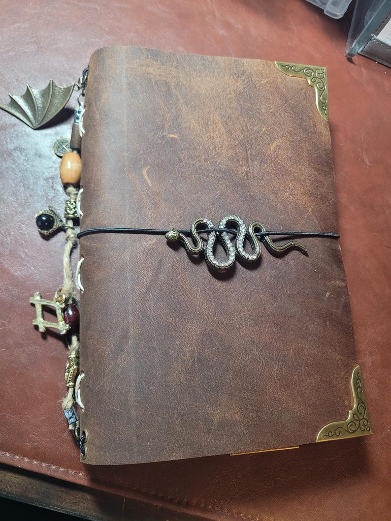 May include: A brown leather journal with decorative metal corner protectors and a snake-shaped clasp. A charm with a bat wing design and other decorative charms hang from the side. The journal is closed with a black elastic band.