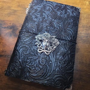 May include: Black leather journal with embossed floral pattern and silver metal corner protectors. The journal features a silver Medusa head clasp and a black elastic band closure. The journal is approximately 8 inches long and 6 inches wide.