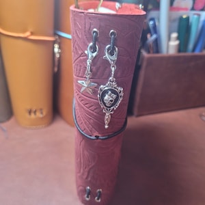 May include: Burgundy leather journal with embossed floral pattern, adorned with silver charms, including a star and a skull design. The journal is secured with a black elastic band and has silver eyelets.