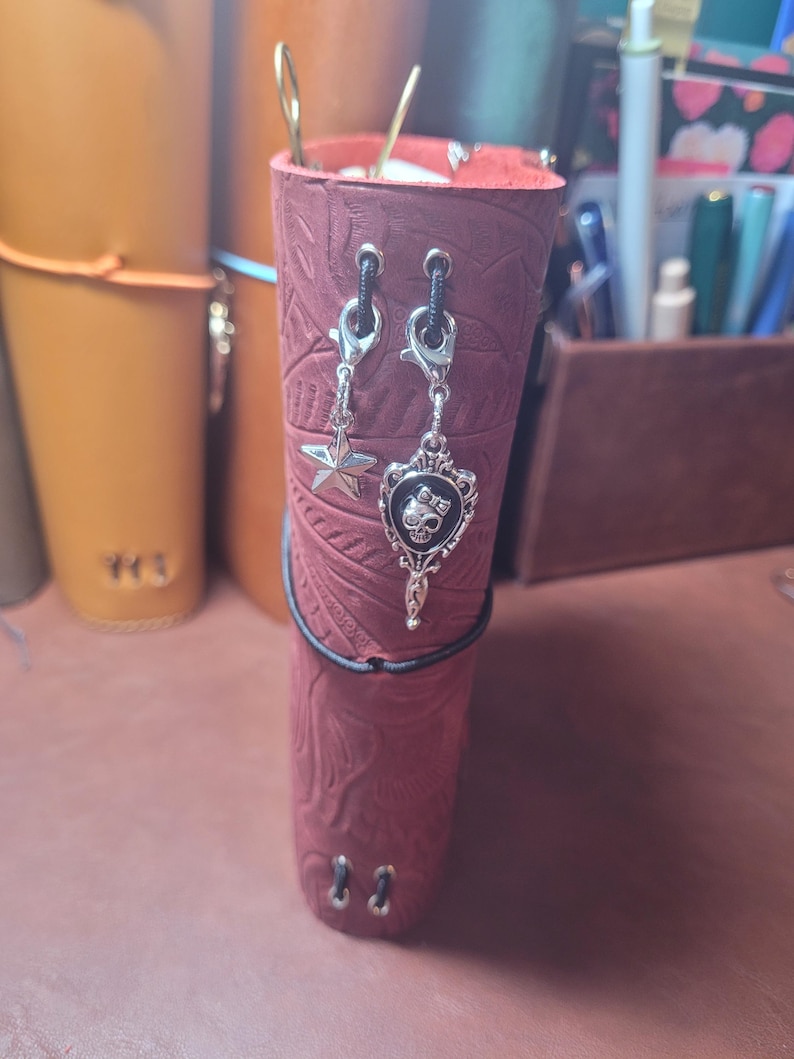 May include: Burgundy leather journal with embossed floral pattern, adorned with silver charms, including a star and a skull design. The journal is secured with a black elastic band and has silver eyelets.