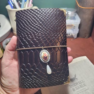 May include: A dark brown leather journal with a reptile skin texture. It features gold corner accents, an elastic closure, and a decorative pendant with a floral design and a pearlescent drop. The journal is held in a hand.
