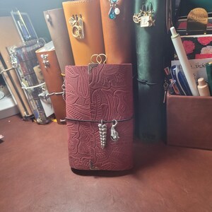 May include: A collection of leather journals in various colors, including burgundy, brown, and green. The burgundy journal in the foreground has a detailed embossed design and decorative charms. Other journals have charms and clasps. The journals are on a brown surface.