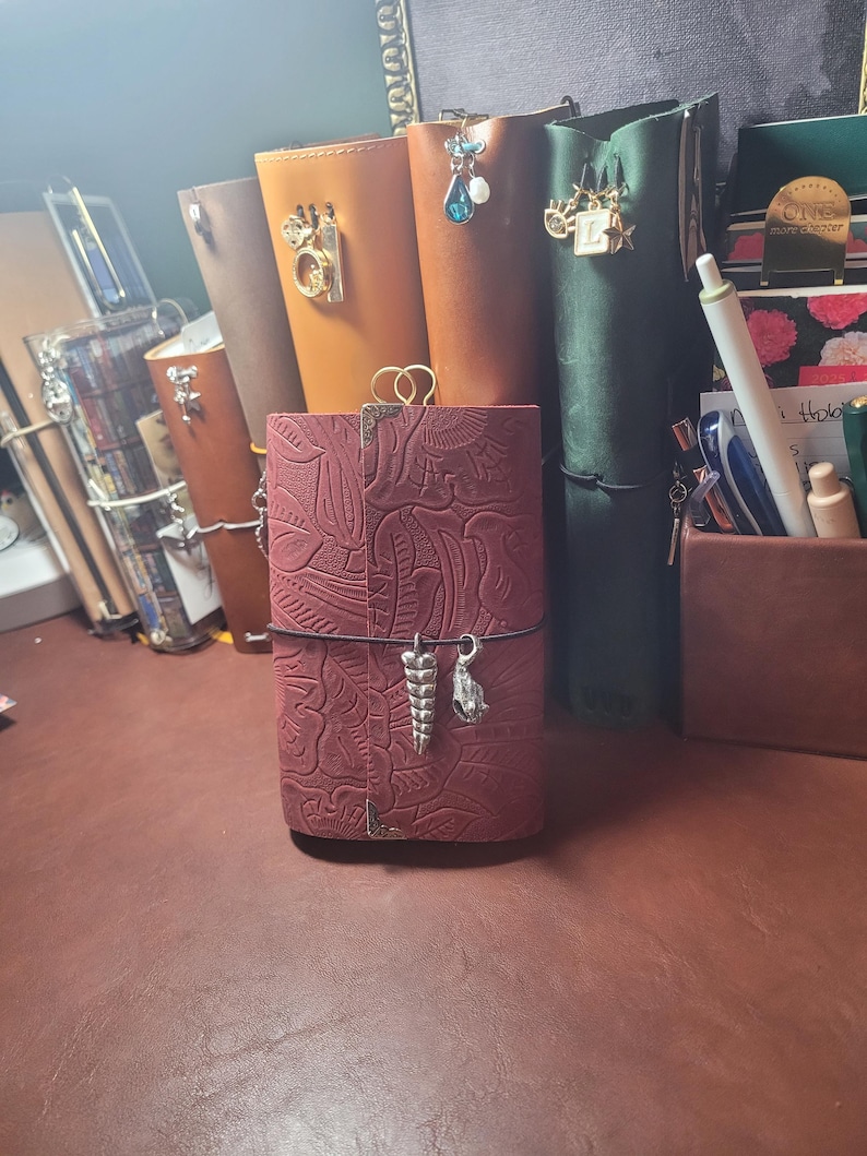May include: A collection of leather journals in various colors, including burgundy, brown, and green. The burgundy journal in the foreground has a detailed embossed design and decorative charms. Other journals have charms and clasps. The journals are on a brown surface.