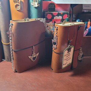 May include: Several handmade leather Traveler's Journals in various colors, adorned with decorative charms. The journals are open, revealing the interior pages. One journal has a tag that reads "Handmade Leather Traveler's Journal".
