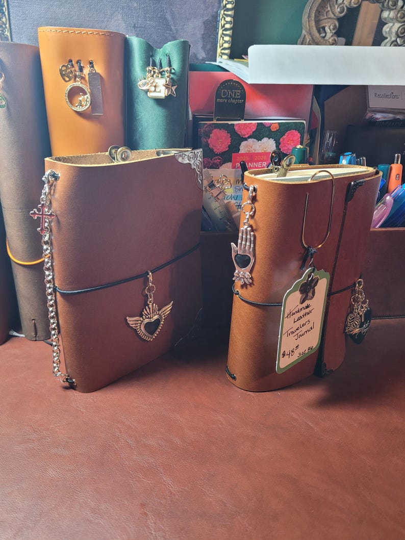 May include: Several handmade leather Traveler's Journals in various colors, adorned with decorative charms. The journals are open, revealing the interior pages. One journal has a tag that reads "Handmade Leather Traveler's Journal".
