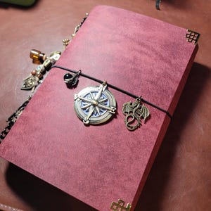 May include: Burgundy-colored journal with decorative corner protectors and a black elastic closure. The journal features a compass charm, a dragon charm, and other decorative charms. The journal is on a brown surface.