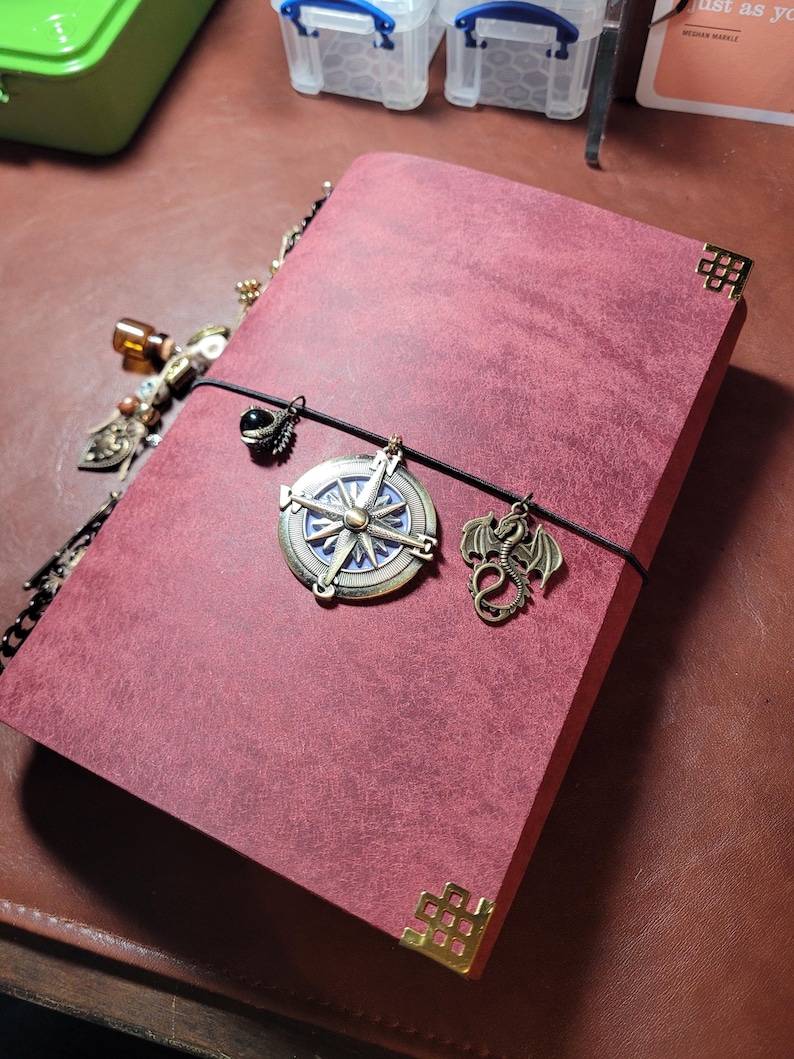 May include: Burgundy-colored journal with decorative corner protectors and a black elastic closure. The journal features a compass charm, a dragon charm, and other decorative charms. The journal is on a brown surface.