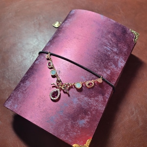 May include: A magenta journal with a decorative gold-toned corner and a black elastic closure. A gold-toned necklace with pink and blue gemstones is draped across the journal. The journal has a textured, marbled appearance.