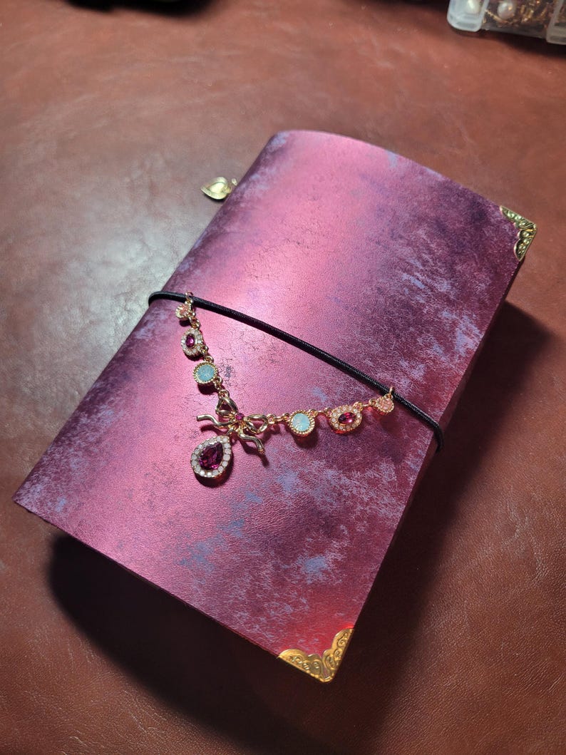May include: A magenta journal with a decorative gold-toned corner and a black elastic closure. A gold-toned necklace with pink and blue gemstones is draped across the journal. The journal has a textured, marbled appearance.