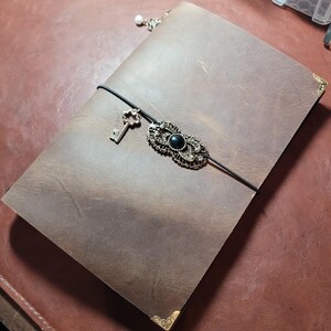 May include: A brown leather journal with a decorative clasp and key charm. The journal has a black elastic closure and gold corner protectors. A small pearl charm hangs from the top. The clasp features a black stone.