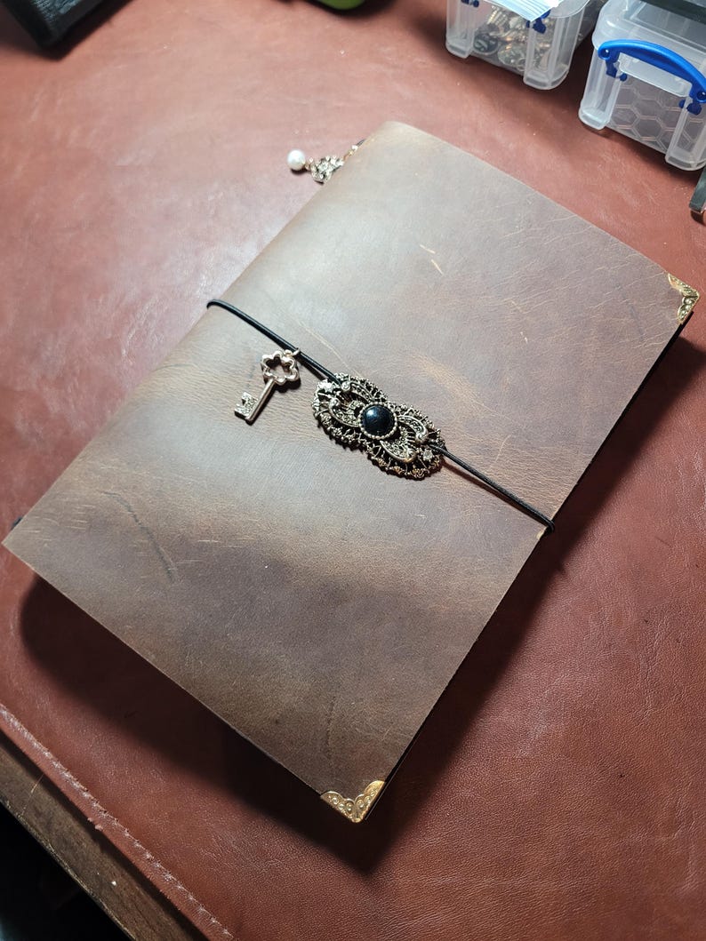 May include: A brown leather journal with a decorative clasp and key charm. The journal has a black elastic closure and gold corner protectors. A small pearl charm hangs from the top. The clasp features a black stone.