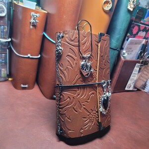 May include: A brown leather journal with embossed floral patterns. The journal is adorned with silver charms and a black elastic closure. Other leather journals in various colors are visible in the background.